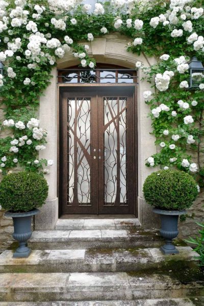 Custom wrought iron front doors with tree branch design, bronze finish double doors, luxury stone entryway with white climbing roses.